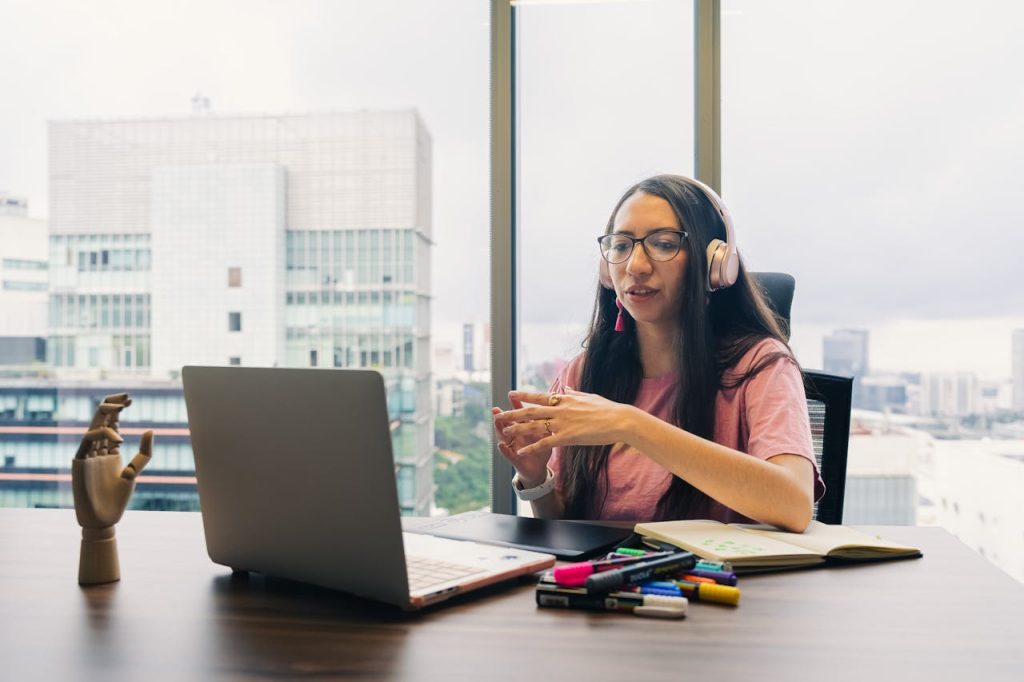 Young woman in pink top conversing on a video call with headphones, in a modern office setting.